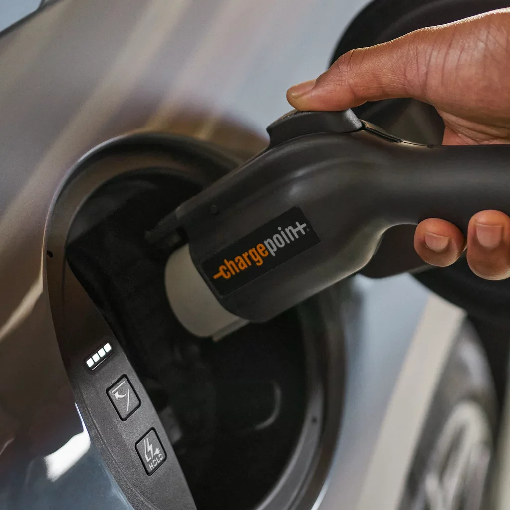 A hand holds a chargepoint+ EV charger, plugging it into a dark gray electric car.