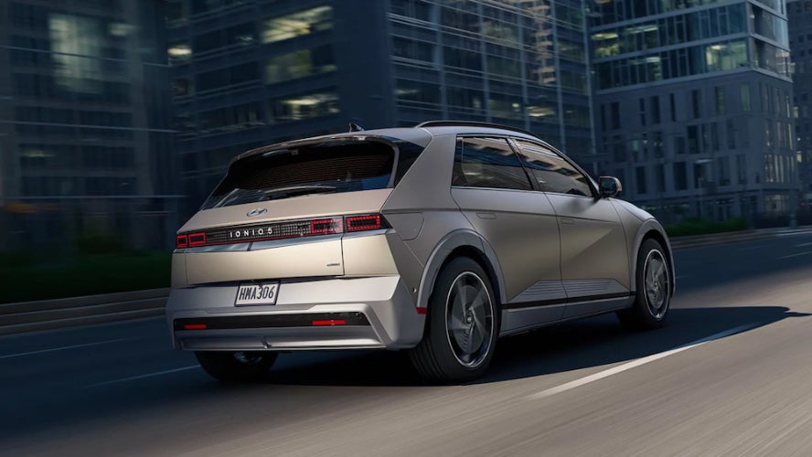A gold 2026 Hyundai Ioniq 5 driving down a city street with glass-fronted buildings lining the road
