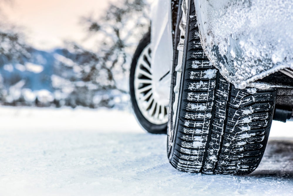 A closeup of a vehicle’s snowy tires, with snow-covered trees and a snowy field in the background