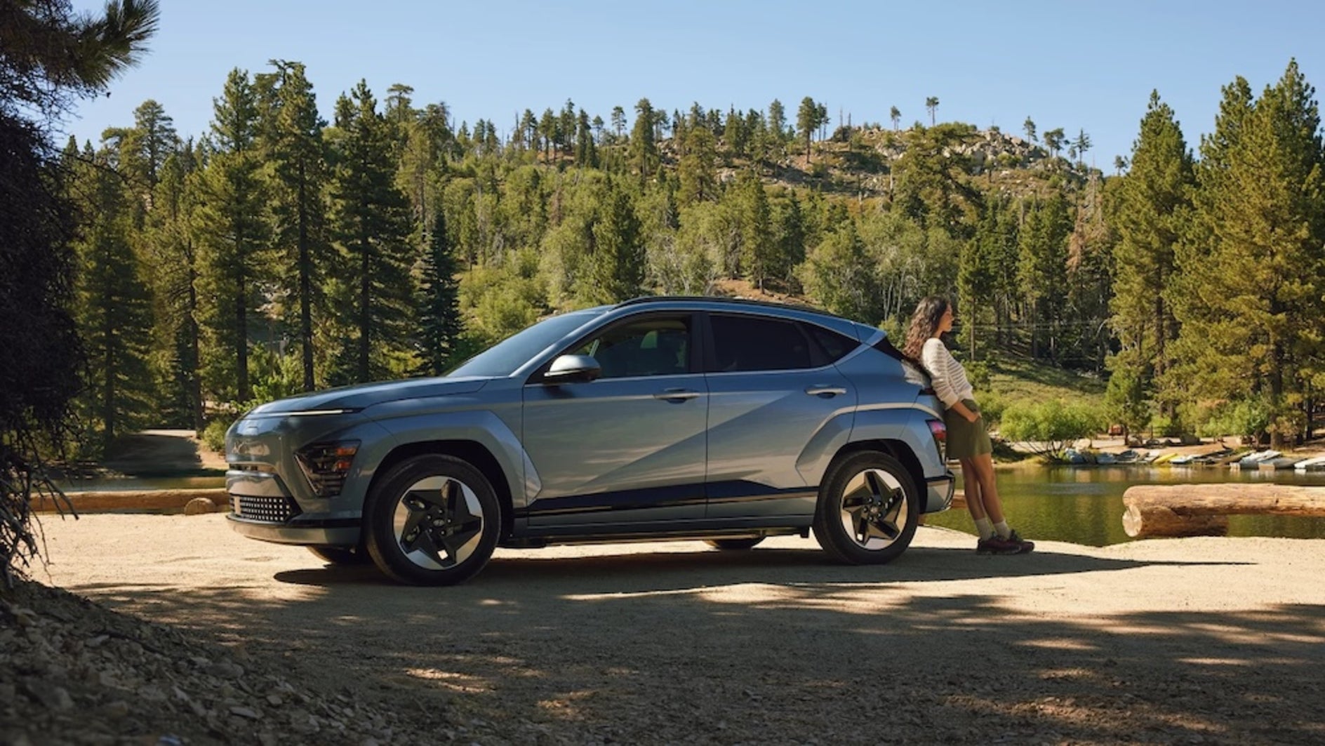 A woman with curly brown hair leaning against the back of a light blue 2026 Hyundai Kona electric, looking out at a lake in the woods