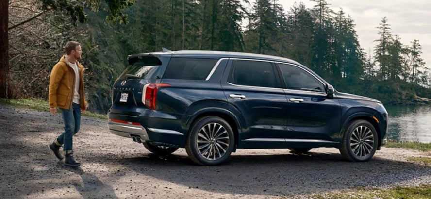 A man in a yellow jacket walking toward a dark blue 2025 Hyundai Palisade SUV parked at the edge of a body of water, with pine trees in the distance