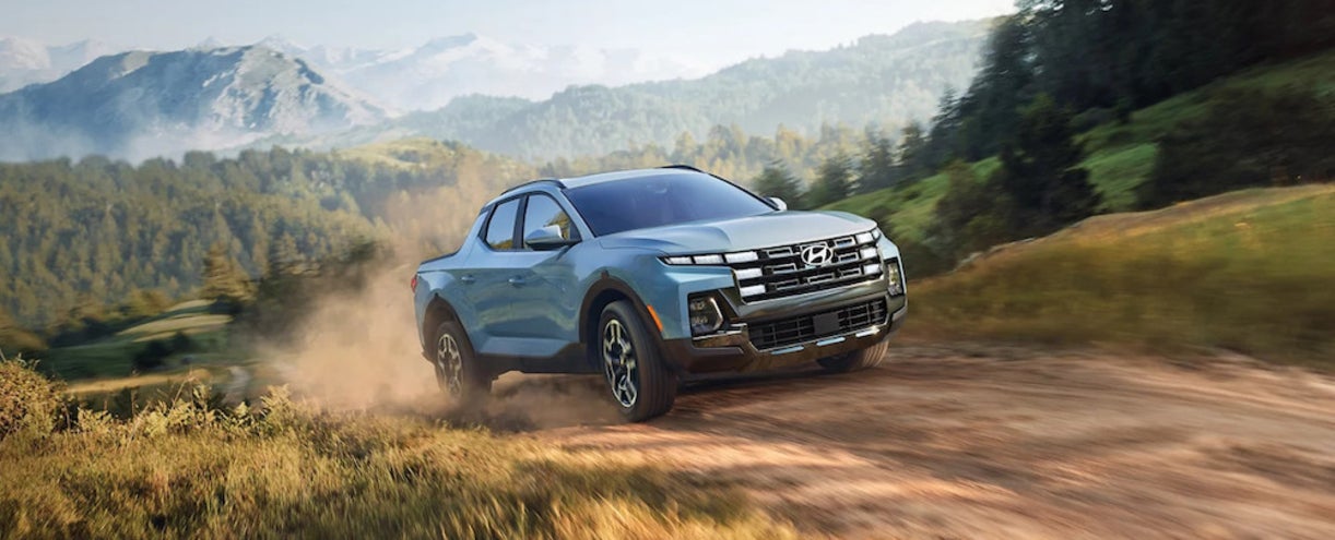 A light blue 2026 Hyundai Santa Cruz driving down a dusty dirt road, with mountains in the background