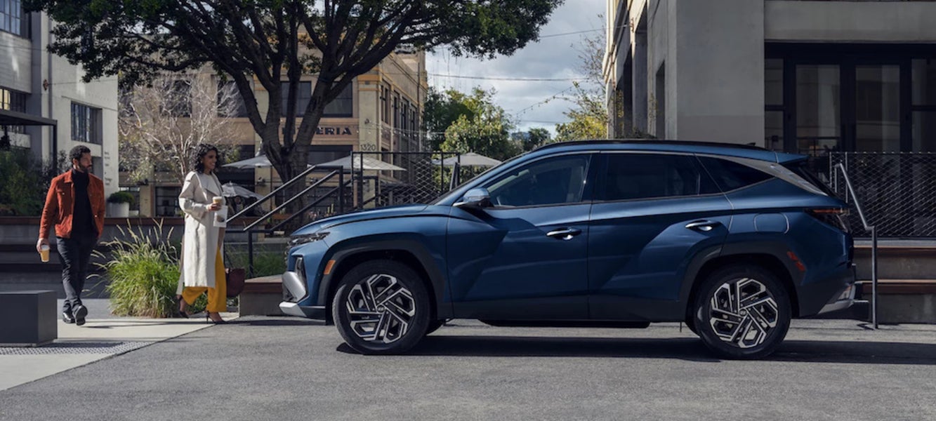 A side view of a blue 2025 Hyundai Tucson in a parking lot, with two people walking toward the SUV